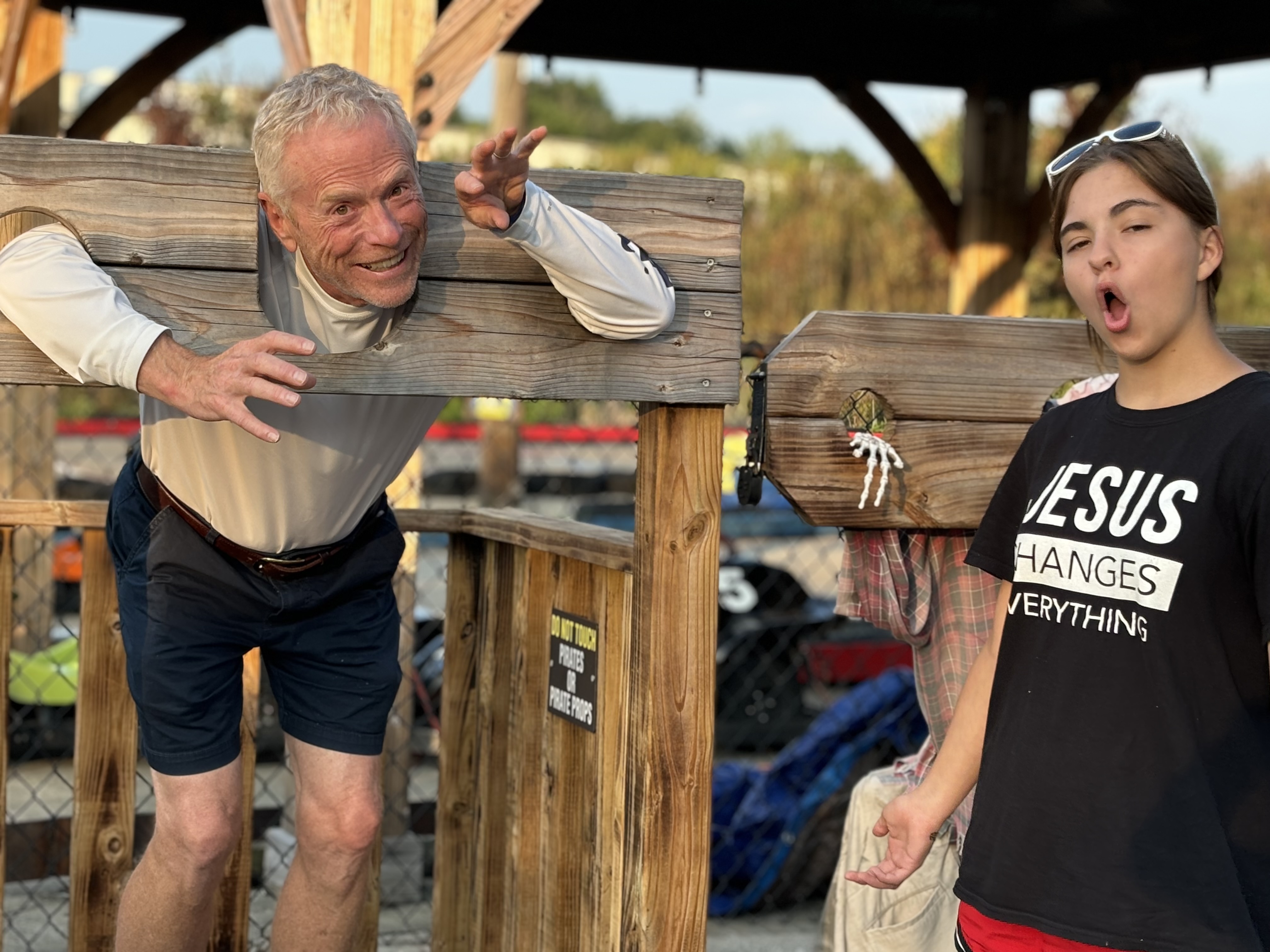 Bob Massie playfully posing in wooden stocks with a young family member nearby, laughing.